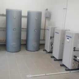 Instalacja kilku zbiorników buforowych i pomp ciepła marki Samsung w pomieszczeniu technicznym, widoczna instalacja rur i skrzynka elektryczna na ścianie.