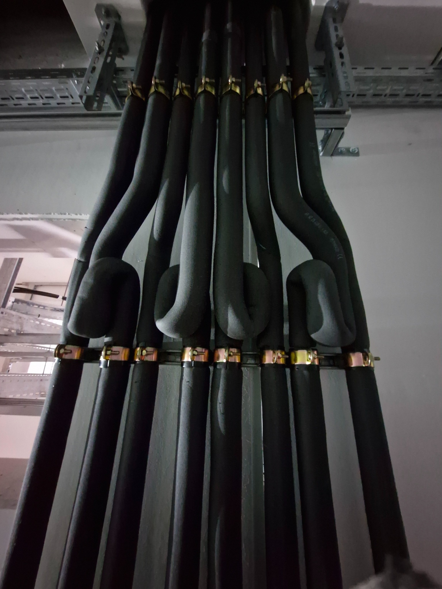 Izolowane rury klimatyzacyjne, zamocowane na metalowej konstrukcji pod sufitem, widoczne złote obejmy mocujące. Instalacja w szarej tonacji.