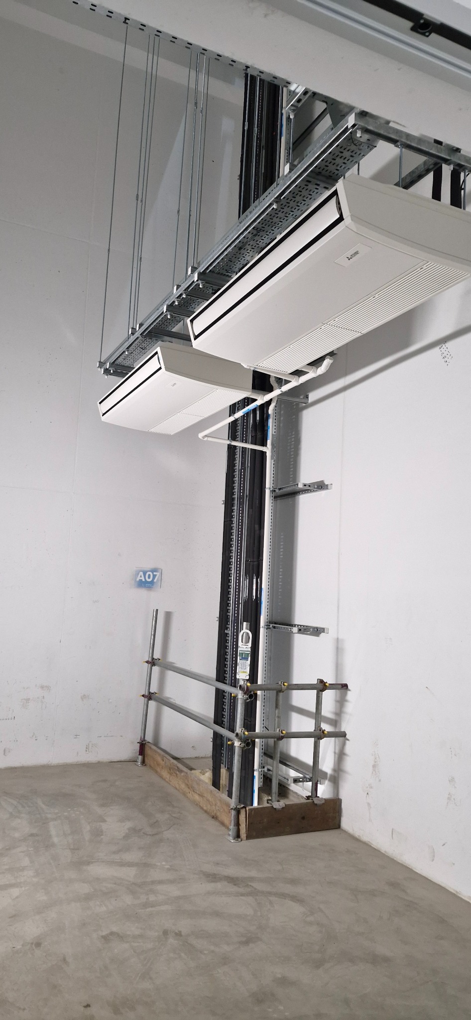 Dwa białe klimatyzatory kasetonowe Mitsubishi zamontowane pod sufitem na metalowej konstrukcji w surowym wnętrzu. Widoczne rury instalacyjne i fragment rusztowania.