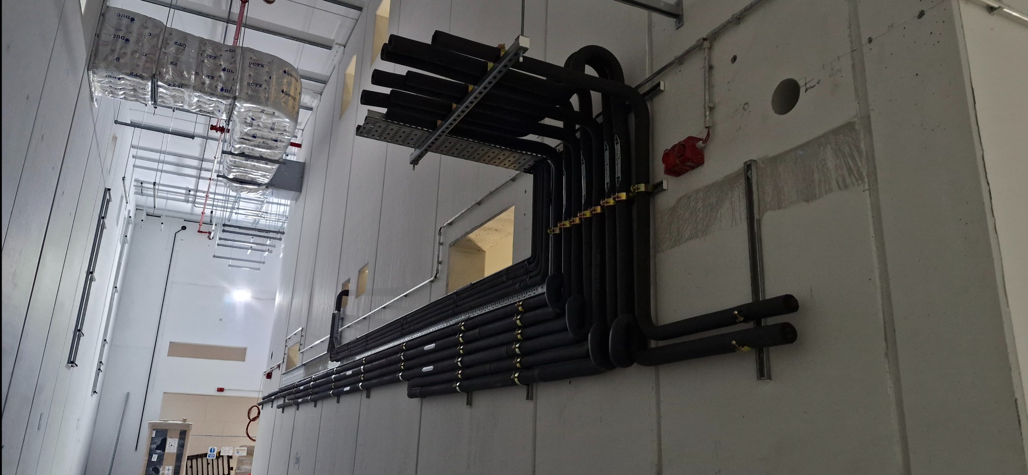 Profesjonalnie poprowadzone, zaizolowane rury instalacji klimatyzacyjnej wzdłuż szarej, betonowej ściany wewnątrz budynku. Widoczne uchwyty mocujące i elementy wentylacji.