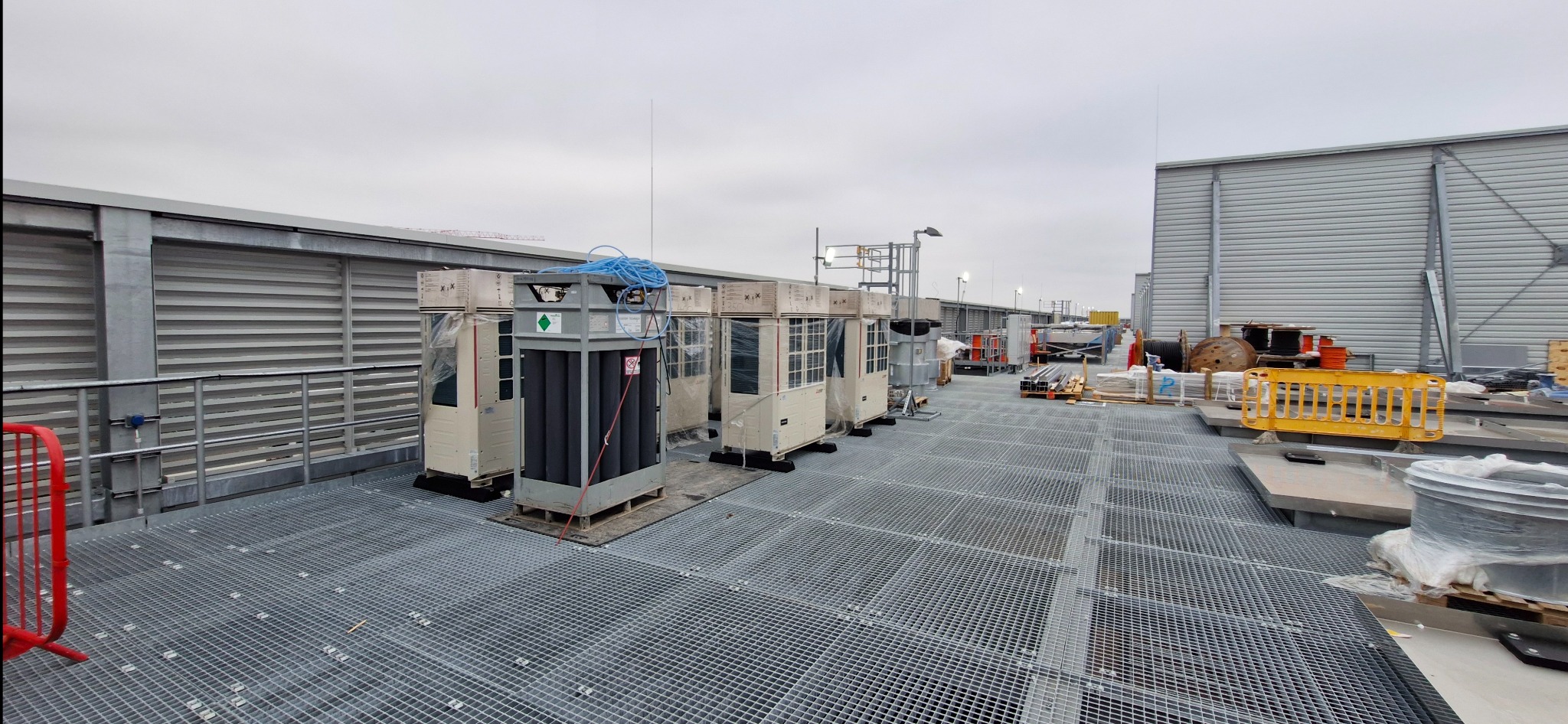 Instalacja klimatyzacji na dachu budynku. Widoczne jednostki zewnętrzne klimatyzatorów, instalacja rurowa i metalowa konstrukcja. Szara, pochmurna pogoda.