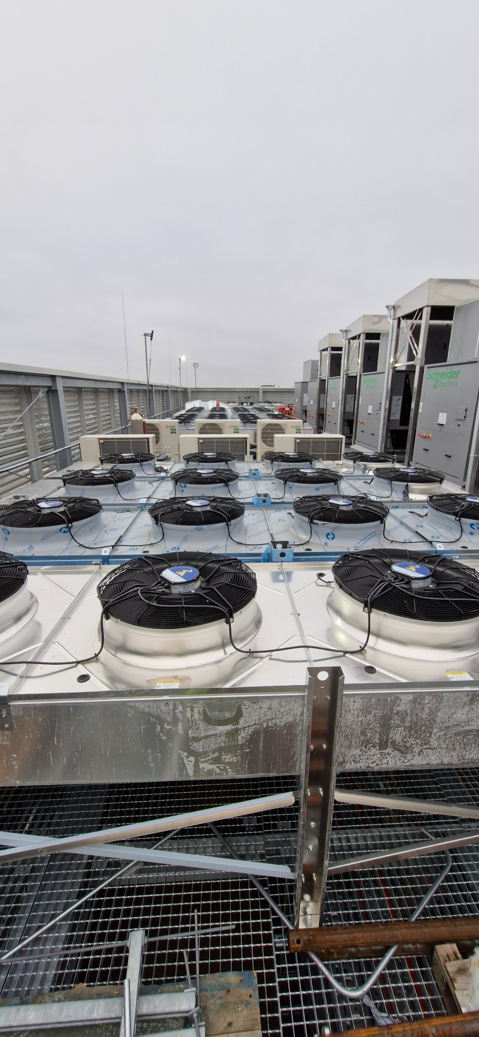 Rozległa instalacja klimatyzacyjna na dachu budynku, z licznymi wentylatorami i jednostkami zewnętrznymi, widocznymi na tle pochmurnego nieba. Metalowa konstrukcja wsporcza.