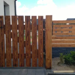 Drewniana furtka wejściowa z pionowych desek, z czarnym uchwytem i widocznymi śrubami mocującymi, przylegająca do ściany budynku i połączona z poziomym ogrodzeniem na betonowej podmurówce.