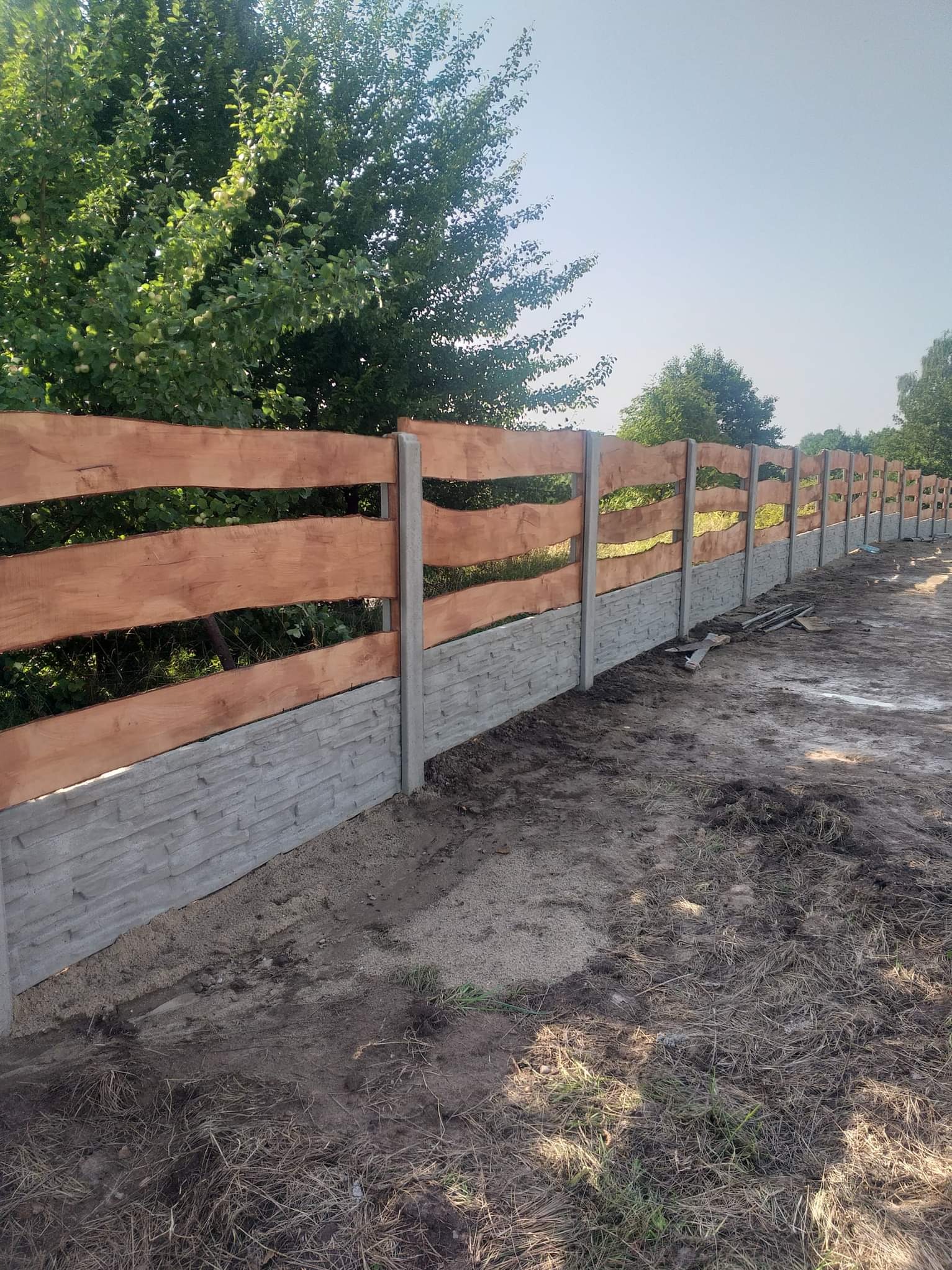 Długie ogrodzenie w stylu ranczo z drewnianymi, falowanymi panelami i betonową podmurówką z fakturą kamienia, ustawione wzdłuż granicy działki z zielonymi drzewami w tle.