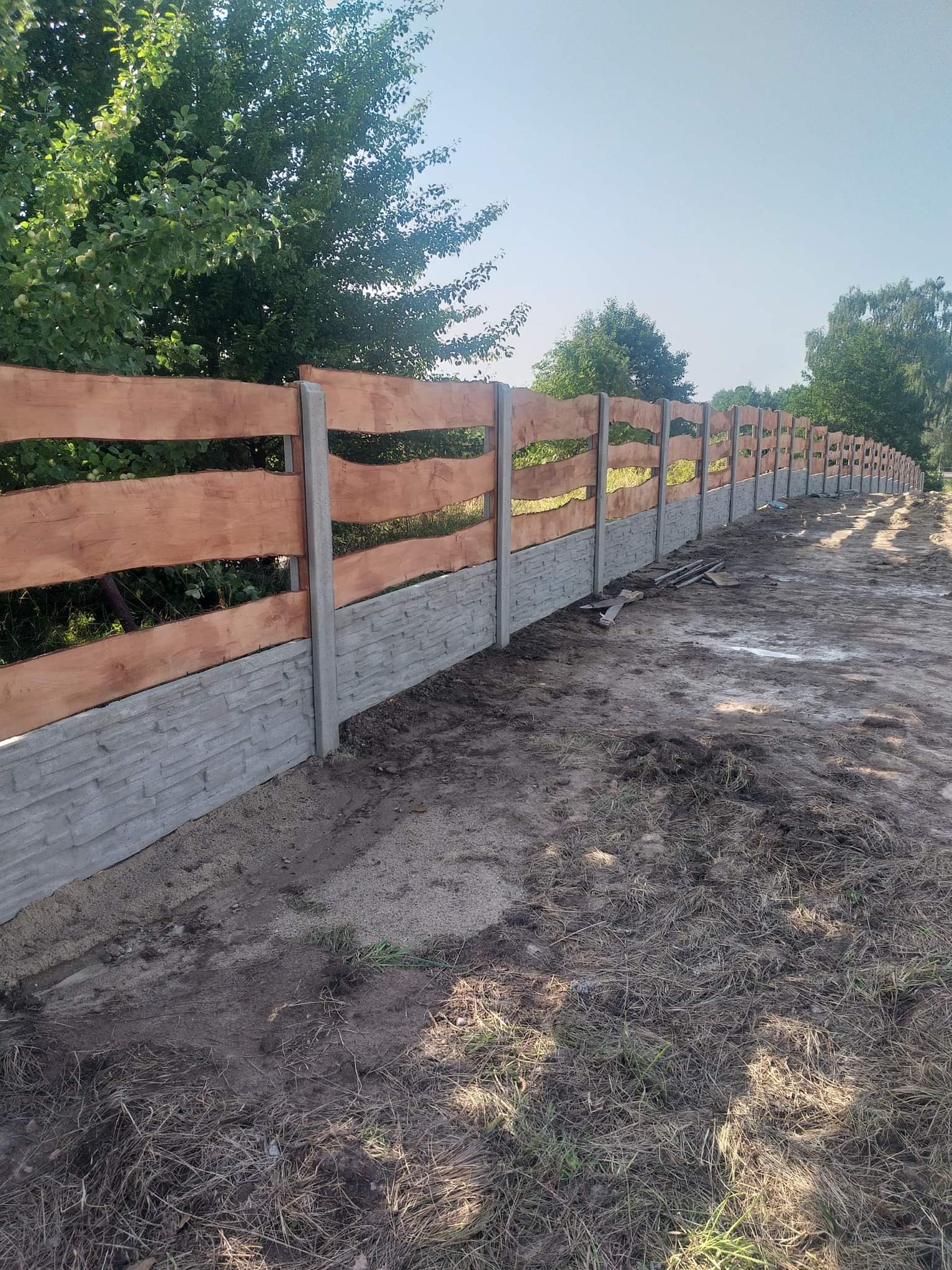 Długi, niski płot w stylu ranczo z poziomych desek w ciepłym odcieniu brązu, osadzony na betonowej podmurówce z fakturą kamienia i słupkach, ciągnący się wzdłuż polnej drogi, z zielenią drzew w tle.