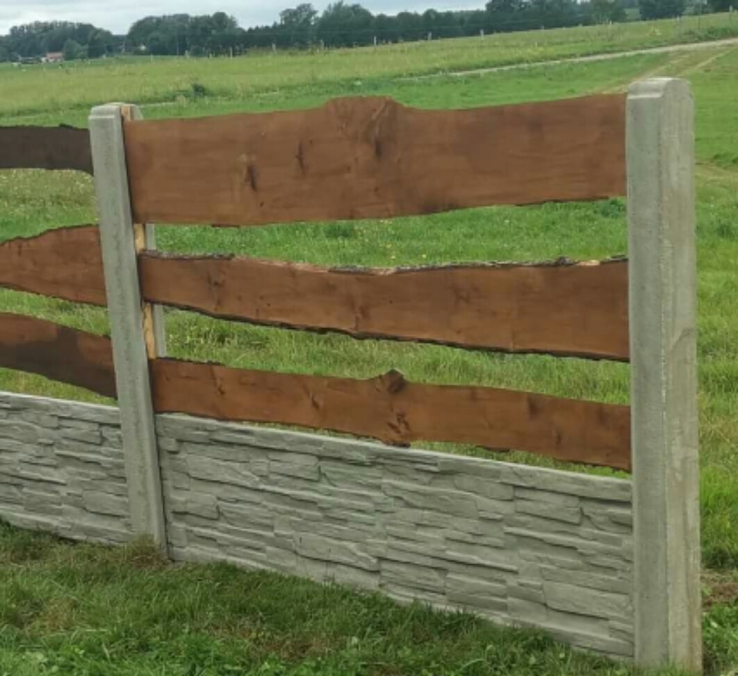 Fragment ogrodzenia w stylu ranczo z drewnianymi, nieregularnymi deskami i betonową podmurówką imitującą kamień, osadzony na betonowych słupkach na tle zielonego pola.
