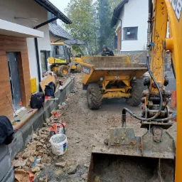 Prace ziemne na budowie: widok z perspektywy koparki JCB na wywrotkę Benford z operatorem w kapturze, w tle żółta ładowarka Mecalac, obok fundamentów domu widoczne narzędzia i materiały budowlane.