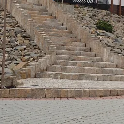 Kamienne schody prowadzące na wzgórze, wykończone kostką brukową, otoczone kamieniami i zielenią. Widoczne różne odcienie szarości i brązu. Detal kostki brukowej na pierwszym planie.