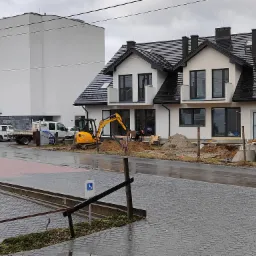 Żółta mini-koparka JCB przy wykopach fundamentowych nowego, dwukondygnacyjnego domu z czarną dachówką na tle nowoczesnego budynku.