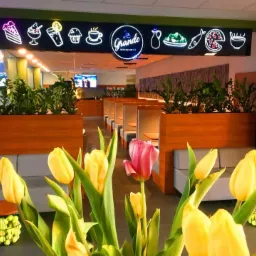 Wnętrze restauracji z neonowym szyldem 'La Grande Restaurant & Co' przedstawiającym ikony potraw, tulipany w wazonie na pierwszym planie, kabiny dla gości w tle.