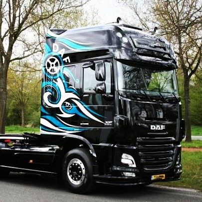 Czarna ciężarówka DAF XF z niestandardową grafiką w kolorach niebieskim i białym na kabinie, przedstawiającą wirujące wzory i logo z kołem, zaparkowana na tle drzew.