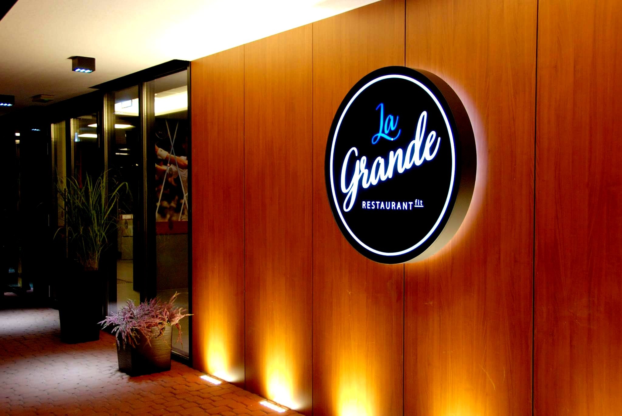 Podświetlany kaseton reklamowy z logo restauracji LaGrande na drewnianej elewacji budynku, oświetlony od dołu punktowymi lampami.