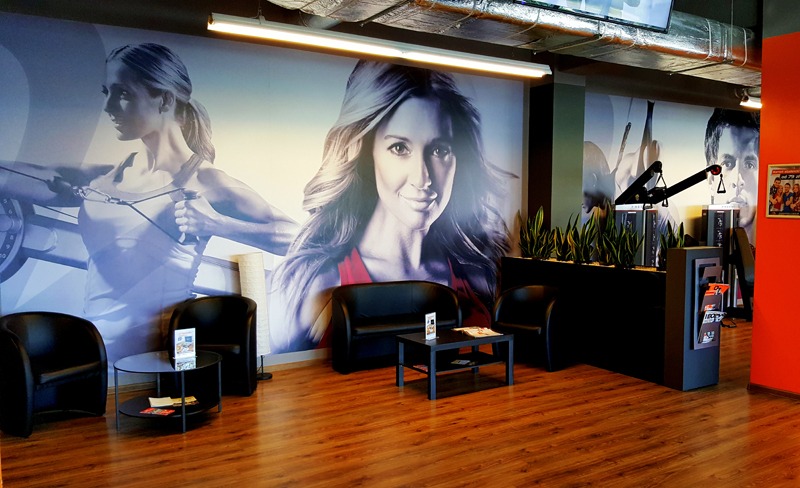 Wnętrze centrum fitness z fototapetą przedstawiającą ćwiczące osoby, czarne skórzane fotele i stoliki kawowe w poczekalni.