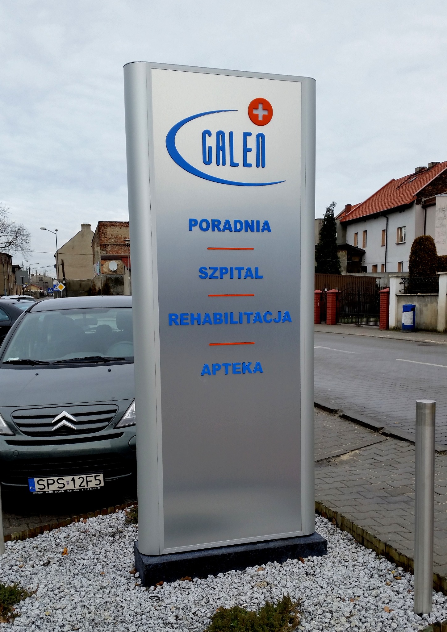 Podświetlany totem reklamowy z logo i informacjami o usługach medycznych firmy Galen: Poradnia, Szpital, Rehabilitacja, Apteka. Totem stoi na żwirowym podłożu, w tle ulica i budynki.