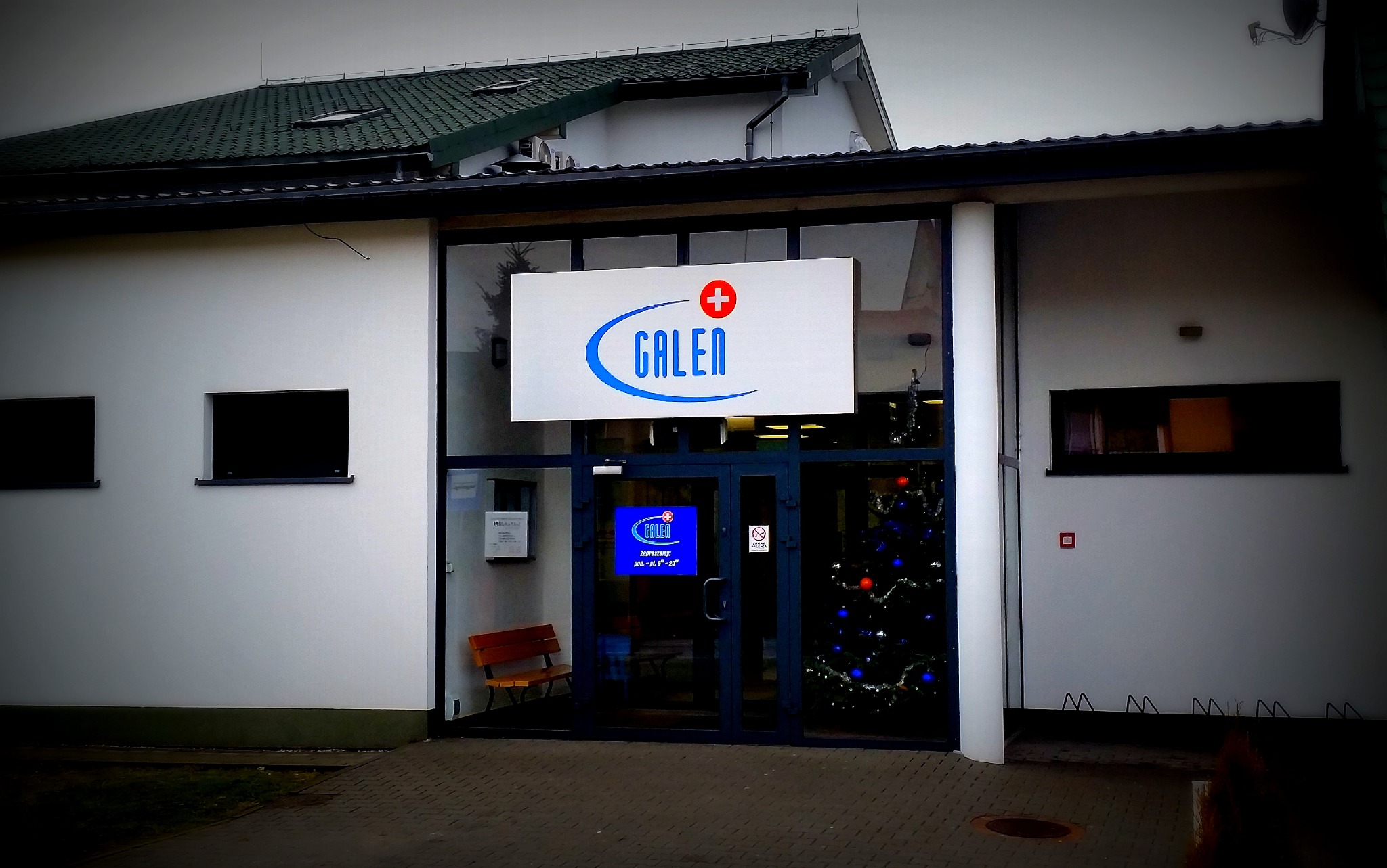 Frezowany, podświetlany kaseton reklamowy firmy Galen nad wejściem do budynku, widoczny fragment elewacji, drzwi wejściowe i świąteczna choinka wewnątrz.