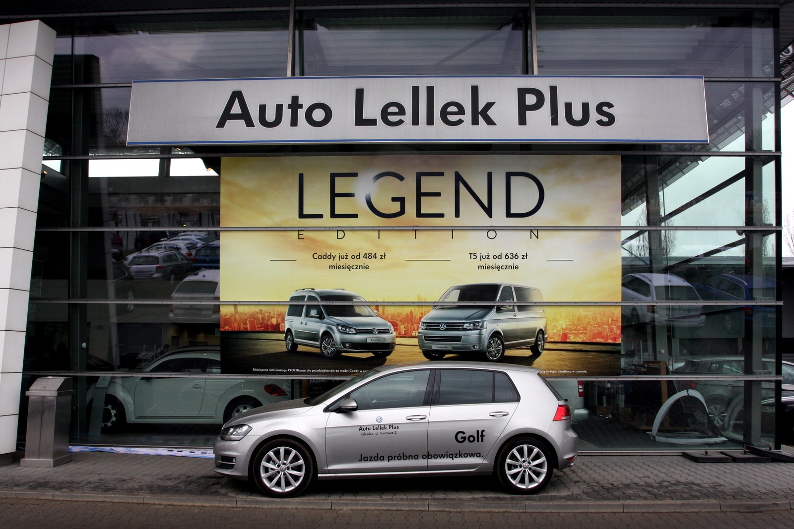 Reklama salonu Auto Lellek Gliwice na witrynie, prezentująca srebrnego Volkswagena Golfa z napisem 'Jazda próbna obowiązkowa' na tle plakatu 'Legend Edition' z modelami Caddy i T5.