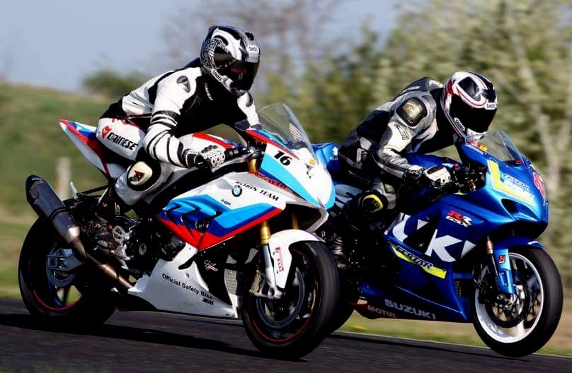 Dwaj motocykliści w kombinezonach i kaskach ścigają się na torze; z przodu biało-czerwono-niebieski motocykl BMW z numerem 16, za nim niebieski motocykl Suzuki.