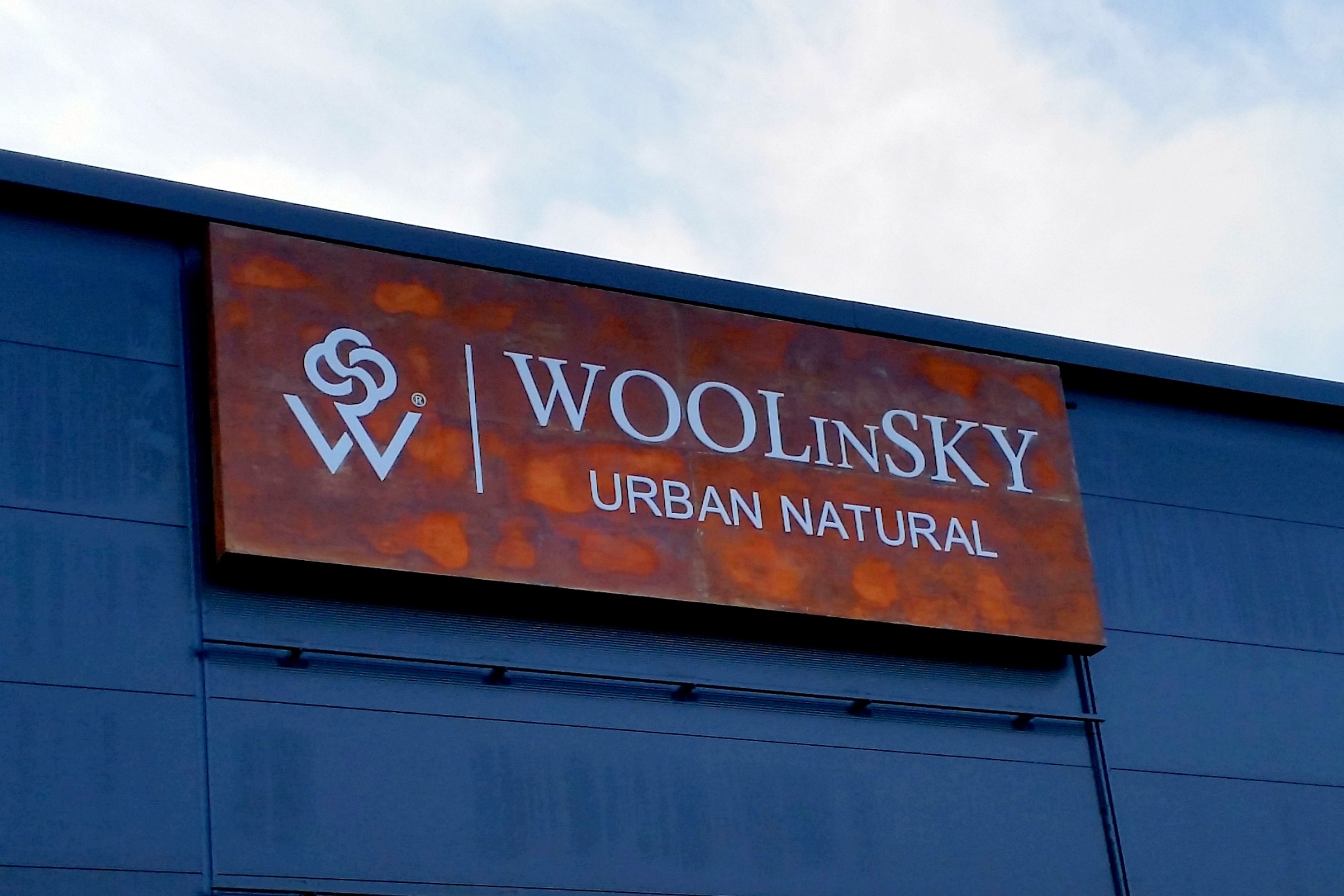 Podświetlany kaseton reklamowy z efektem cortenu, z logo i nazwą firmy Wolinsky, zamontowany na elewacji budynku.