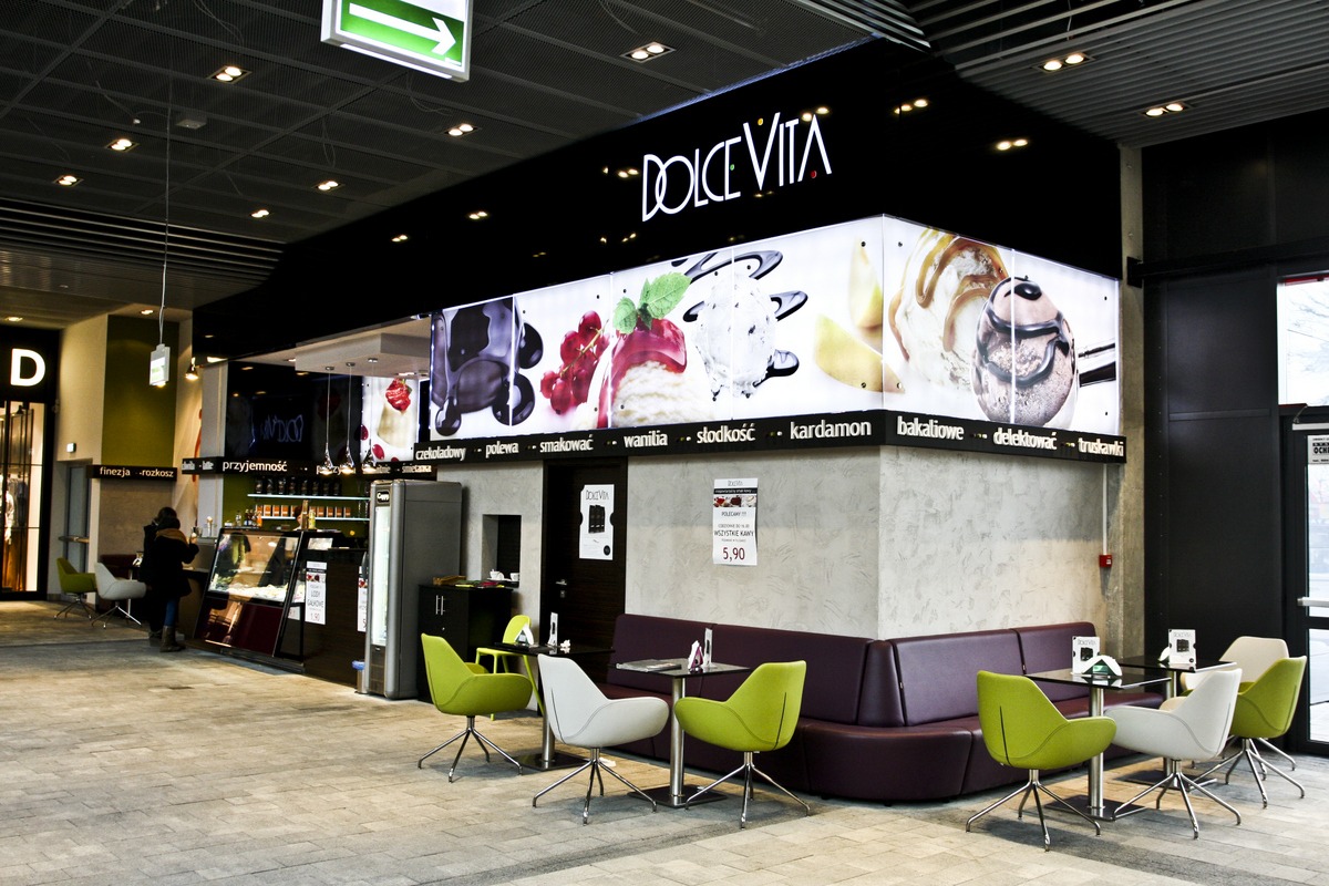 Wnętrze kawiarni Dolce Vita z podświetlanym panelem reklamowym lodów i deserów, zielonymi krzesłami i ciemnofioletową sofą narożną.