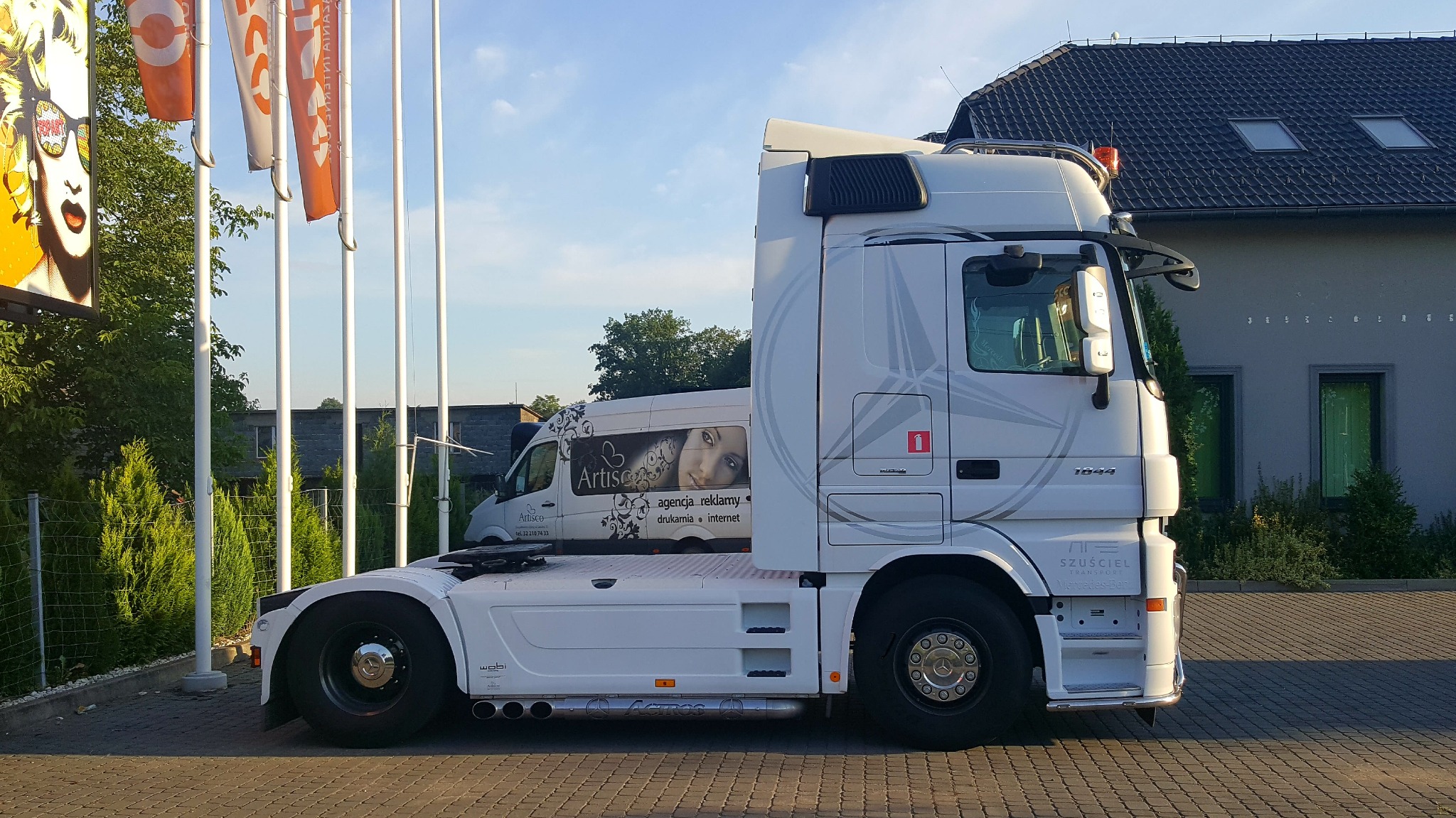 Biała ciężarówka z naczepą, z grafiką reklamową na kabinie, zaparkowana na brukowanym placu, w tle budynek z ciemnym dachem i reklama wizualna na słupach.
