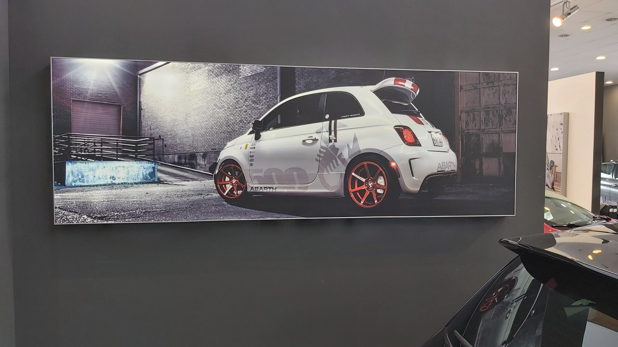 Duży, panoramiczny wydruk na ścianie przedstawia biały samochód Abarth z pomarańczowymi felgami na tle miejskiego krajobrazu z garażem i ceglanymi ścianami. Wydruk wisi w salonie samochodowym.