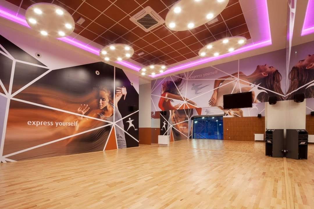 Wnętrze sali fitness z fototapetami przedstawiającymi dynamiczne sylwetki, podświetlony sufit z okrągłymi lampami, drewniana podłoga.