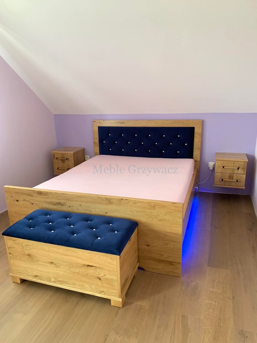 Drewniane łóżko z tapicerowanym, granatowym zagłówkiem z guzikami, uzupełnione skrzynią-pufą w tym samym stylu, podświetlone niebieskim światłem LED, w pokoju na poddaszu.