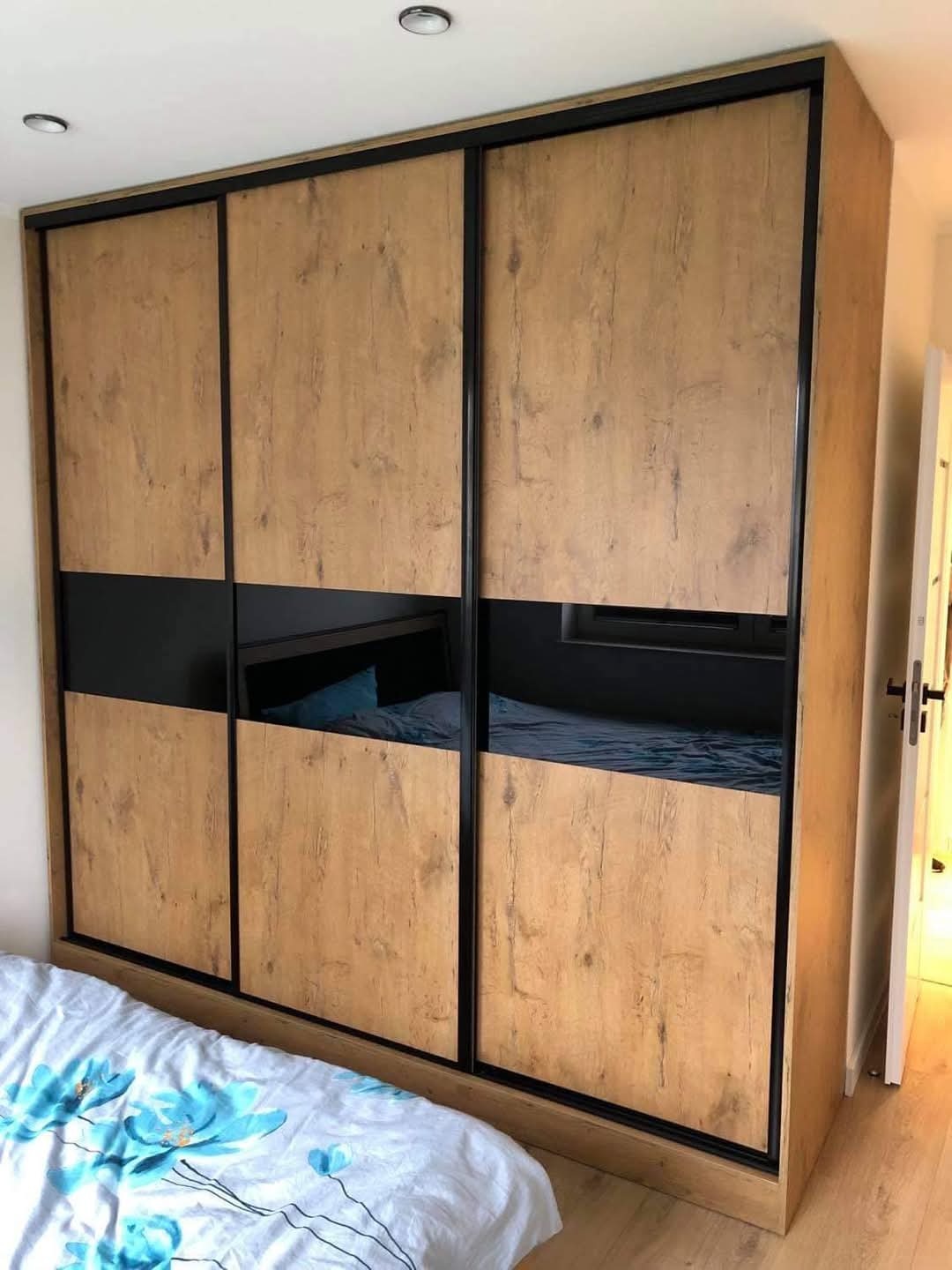 Trzydrzwiowa szafa przesuwna z okleiną imitującą drewno i czarnymi, lustrzanymi wstawkami, widoczna częściowo pościel w kwiatowy wzór.