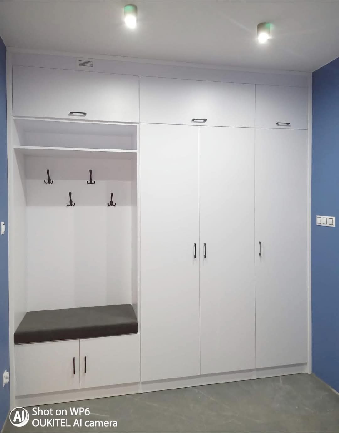 Biała zabudowa meblowa z siedziskiem i wieszakami w przedpokoju, minimalistyczny design, niebieska ściana w tle.