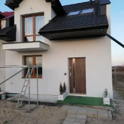 Nowoczesny dom z białą elewacją, ciemną dachówką i drewnianą stolarką okienną, widok z zewnątrz na częściowo zagospodarowany teren przed budynkiem, z widocznym rusztowaniem i elementami...