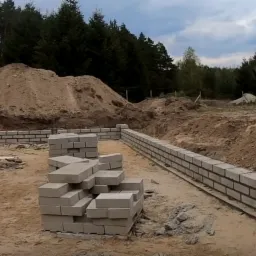 Wznoszenie fundamentów z bloczków betonowych na piaszczystym gruncie, widoczny wykop i pryzma ziemi w tle lasu.