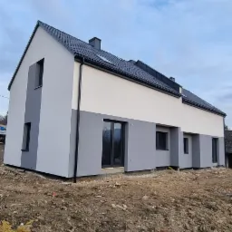 Nowoczesny dom w zabudowie szeregowej z dwukolorową elewacją: szary dół i kremowa góra, czarna dachówka i okna, teren wokół domu jeszcze niezagospodarowany.