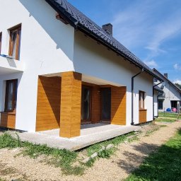 GRYGNY-FIRMA BUDOWLANA - Nowoczesny dom jednorodzinny z białymi ścianami i drewnopodobną okładziną elewacyjną, ciemna dachówka, jasne okna, widok w słoneczny dzień.