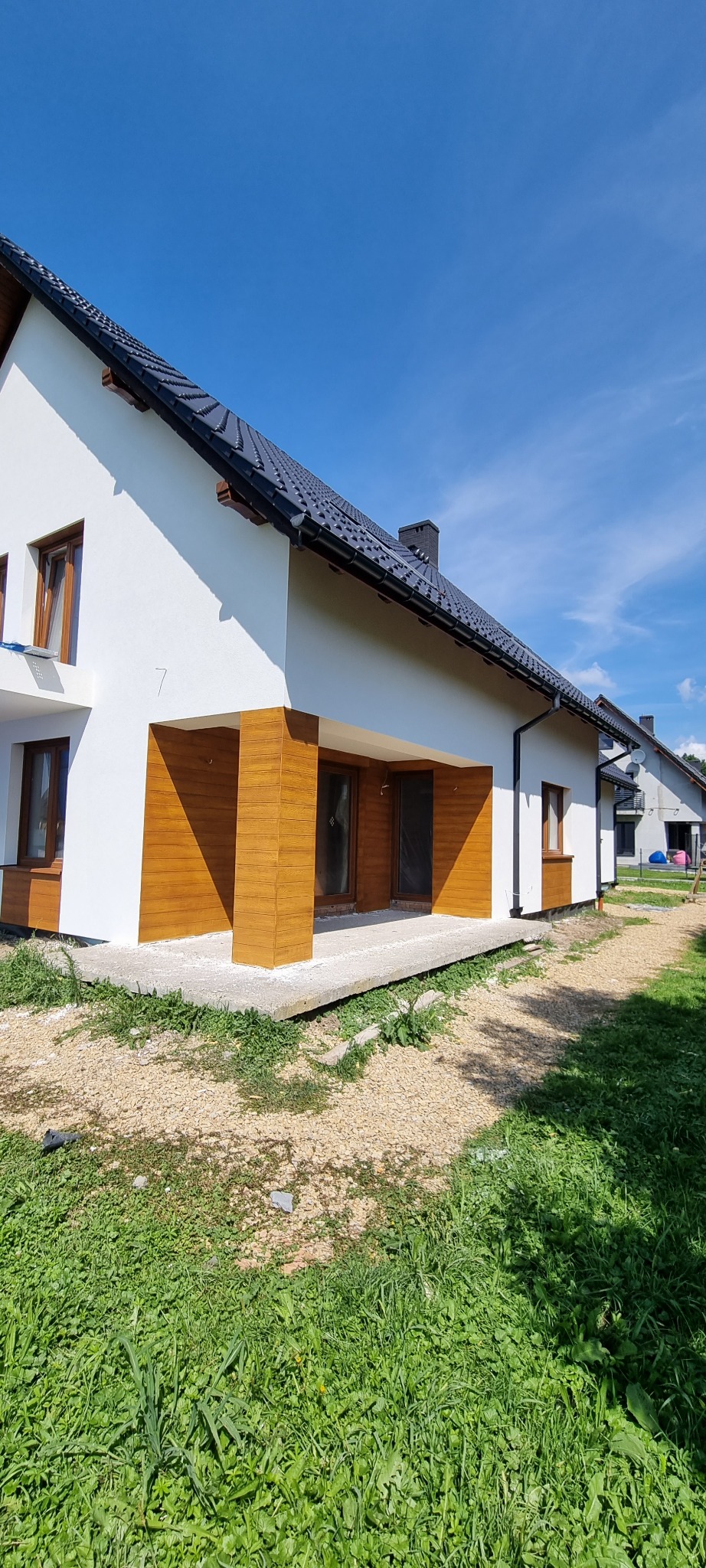 Nowoczesny dom jednorodzinny z białymi ścianami i drewnopodobną okładziną elewacyjną, ciemna dachówka, jasne okna, widok w słoneczny dzień.