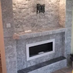 Obudowa kominka elektrycznego wykończona szarą, kamienną okładziną, z zamontowanym uchwytem na telewizor na ścianie, oświetlona od góry.