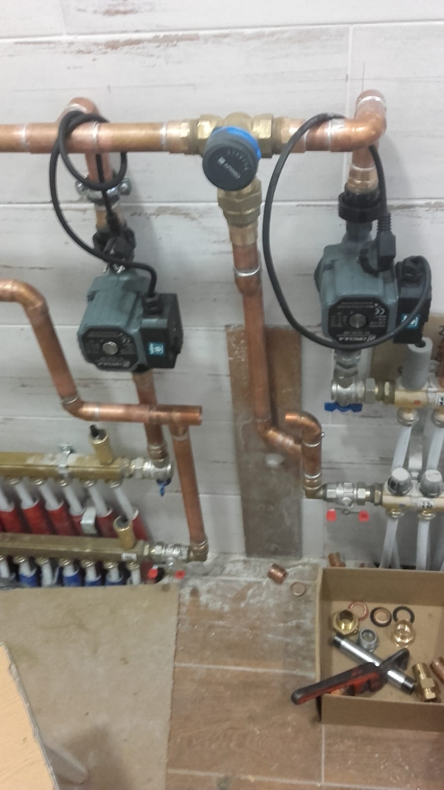 Instalacja centralnego ogrzewania w trakcie montażu: miedziane rury, pompy obiegowe z podłączeniem elektrycznym, rozdzielacz z licznymi zaworami, skrzynka z narzędziami i złączkami na podłodze.