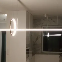 Nowoczesna lampa wisząca LED z okrągłym i liniowym elementem, zamontowana w minimalistycznej kuchni z białymi szafkami i marmurowym backsplashiem, widoczny fragment ściany w trakcie remontu.