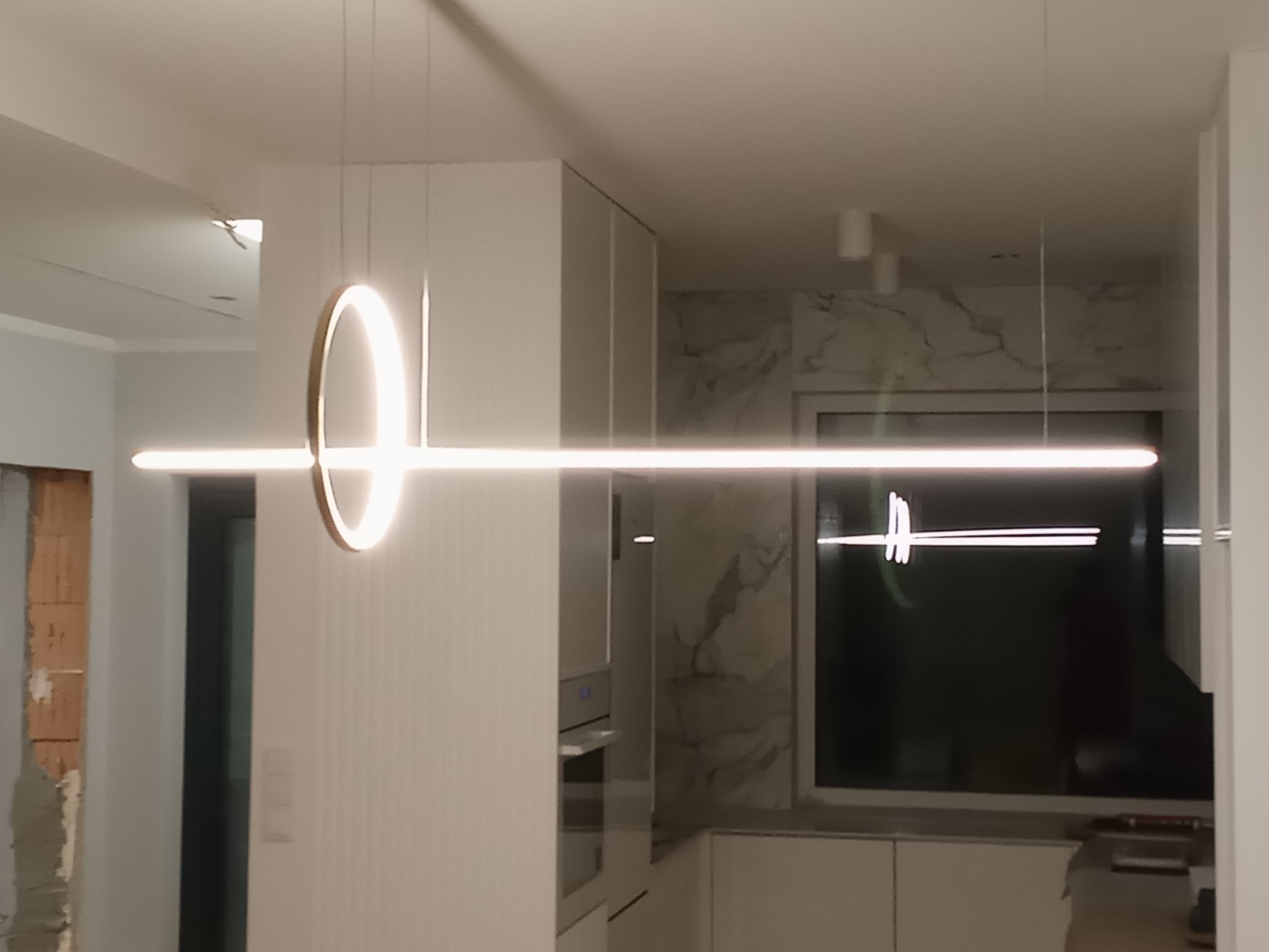 Nowoczesna lampa wisząca LED z okrągłym i liniowym elementem, zamontowana w minimalistycznej kuchni z białymi szafkami i marmurowym backsplashiem, widoczny fragment ściany w trakcie remontu.