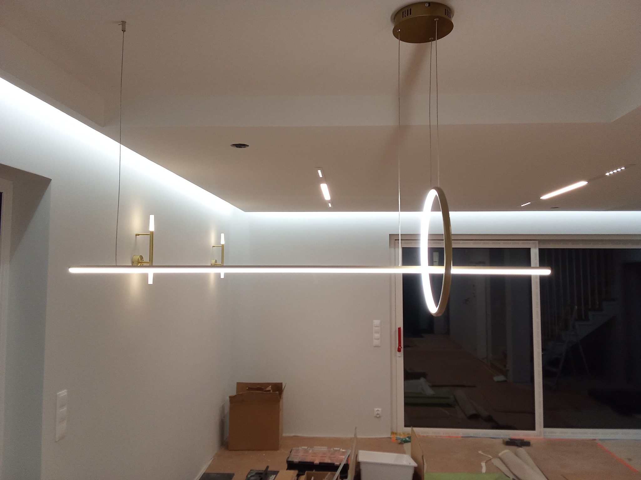 Nowoczesne oświetlenie LED: wisząca lampa pierścieniowa i listwy sufitowe w surowym wnętrzu, widoczne narzędzia i kartony na podłodze sugerują trwający montaż.