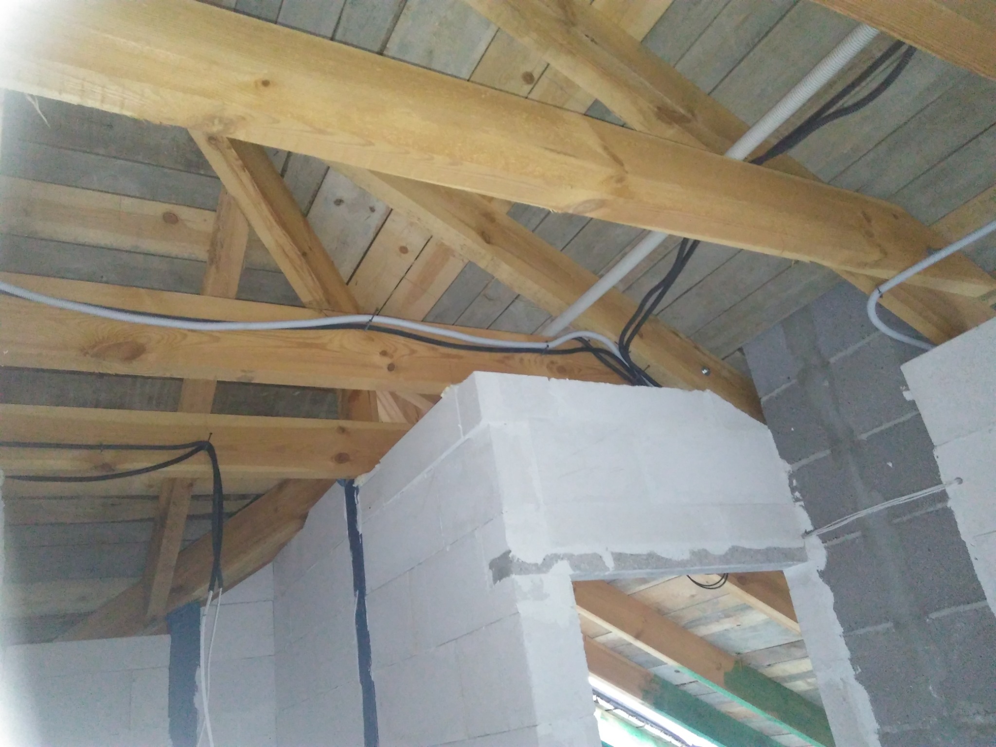Kable elektryczne prowadzone wzdłuż drewnianej konstrukcji dachu, widoczne białe rury karbowane i czarne przewody, ściany z betonu komórkowego w surowym stanie.