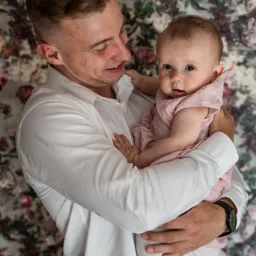 Uśmiechnięty mężczyzna w białej koszuli trzyma na rękach niemowlę w różowej sukience z dekoracyjnym tłem w kwiaty.