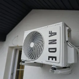 Zewnętrzna jednostka klimatyzacji marki ANDE zamontowana na ścianie budynku z widocznymi przewodami i wspornikami montażowymi, z fragmentem okna i dachu w tle.