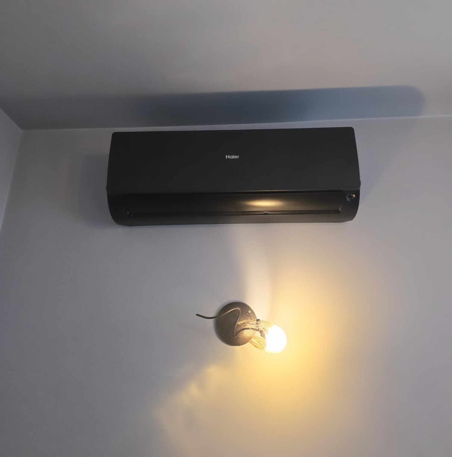 Czarny klimatyzator Haier zamontowany na jasnoszarej ścianie, poniżej widoczna lampka ścienna z żarówką.