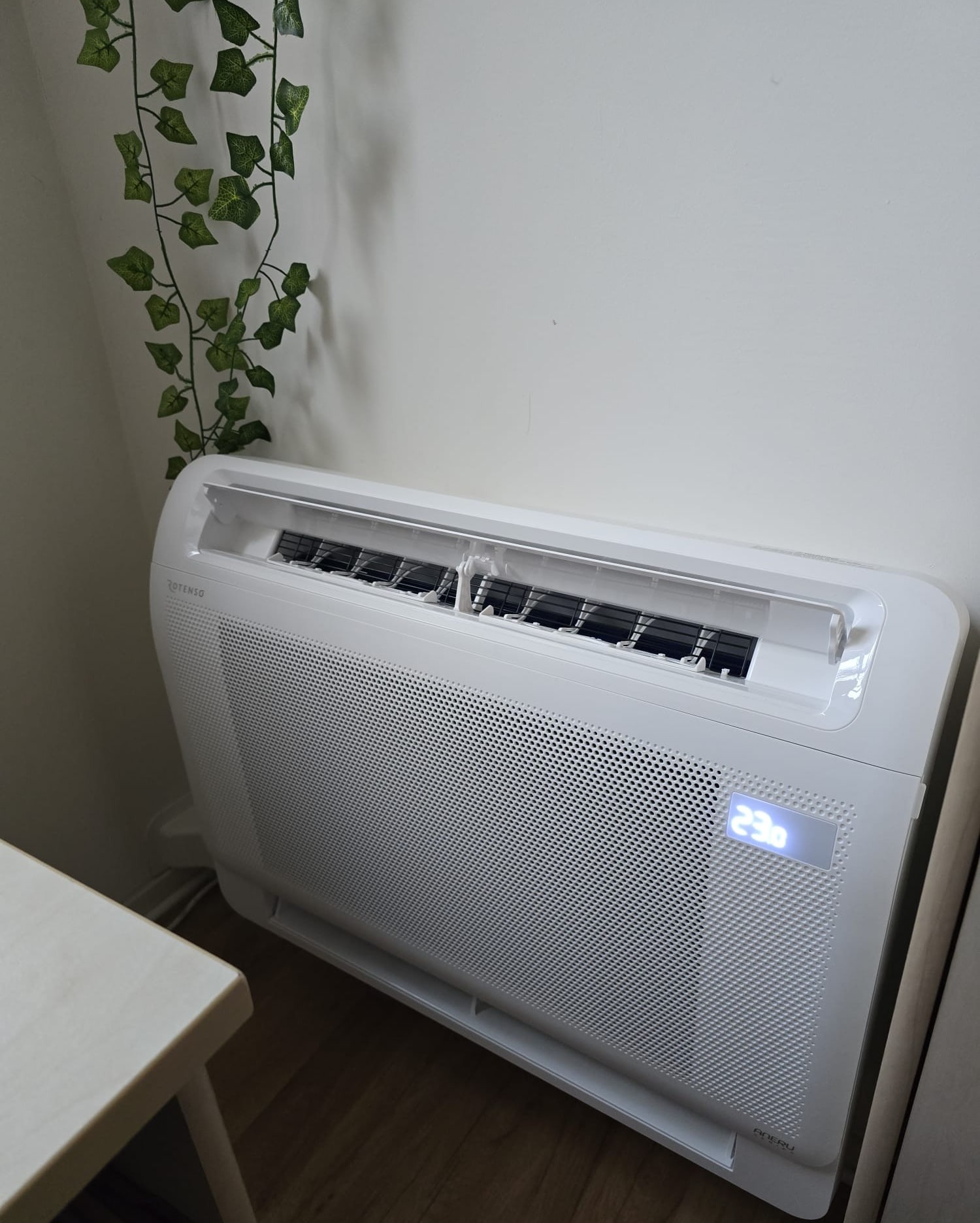 Biała, nowoczesna klimatyzacja podłogowa marki Rotenso z wyświetlaczem temperatury 23 stopnie Celsjusza, zamontowana przy ścianie z dekoracyjną girlandą z bluszczu.