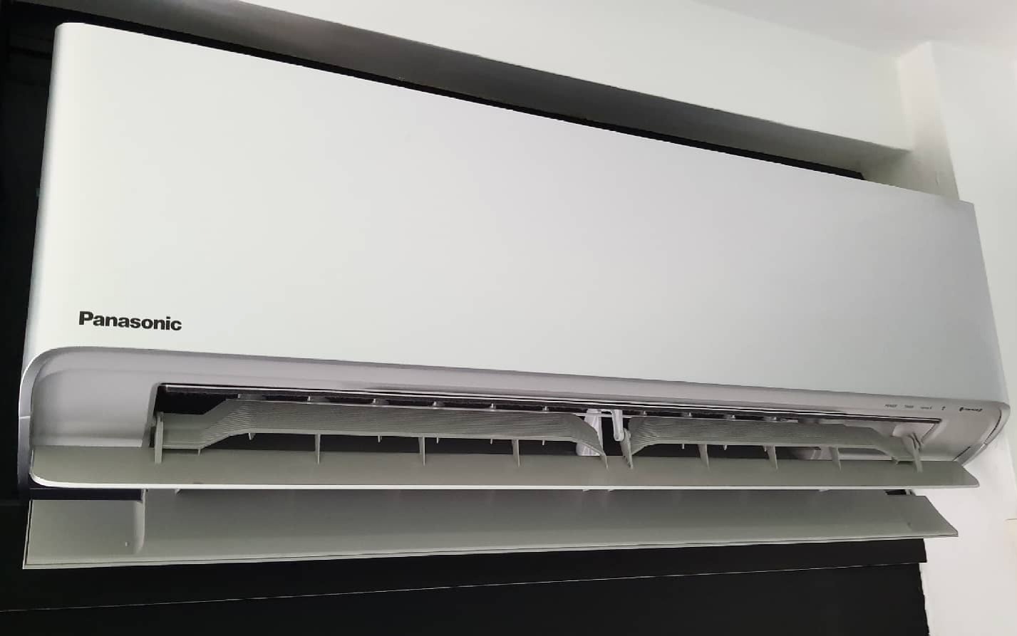 Zamontowana, biała klimatyzacja Panasonic z otwartym panelem, widoczne żaluzje wylotu powietrza, na tle białej ściany i czarnej rolety.