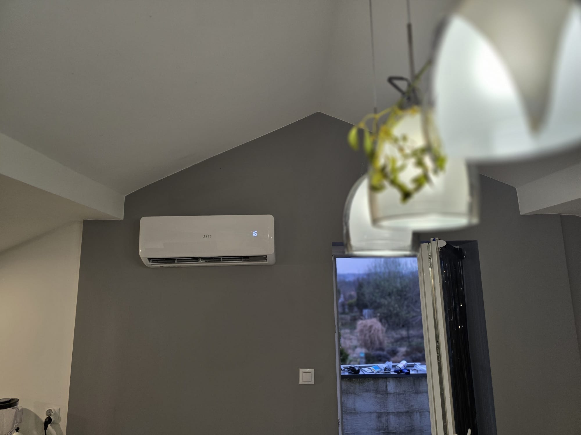 Biały klimatyzator ścienny z wyświetlaczem temperatury (16 stopni) zamontowany na szarej ścianie w pomieszczeniu z widoczną lampą wiszącą i oknem balkonowym.