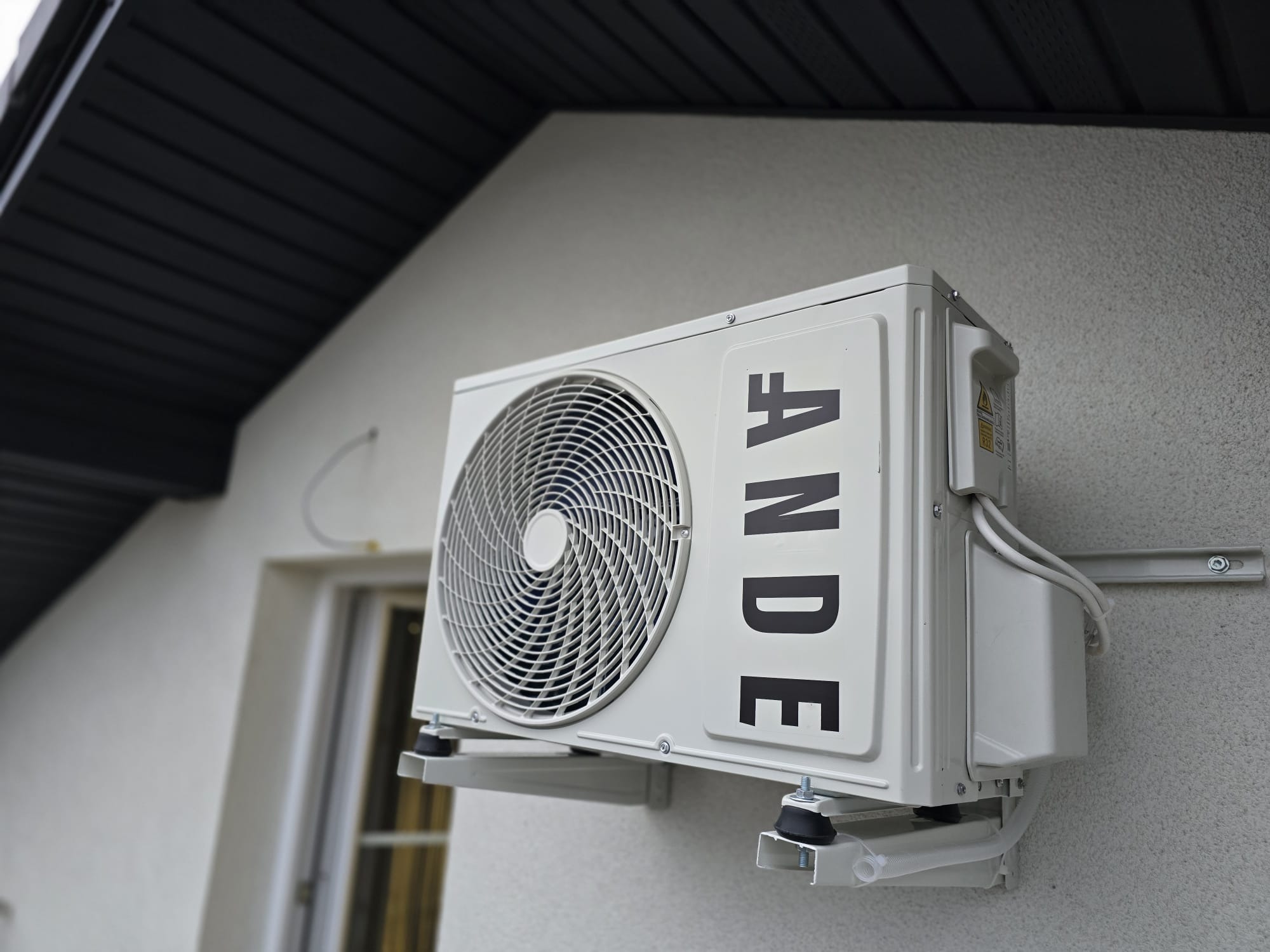 Zewnętrzna jednostka klimatyzacji marki ANDE zamontowana na ścianie budynku z widocznymi przewodami i wspornikami montażowymi, z fragmentem okna i dachu w tle.