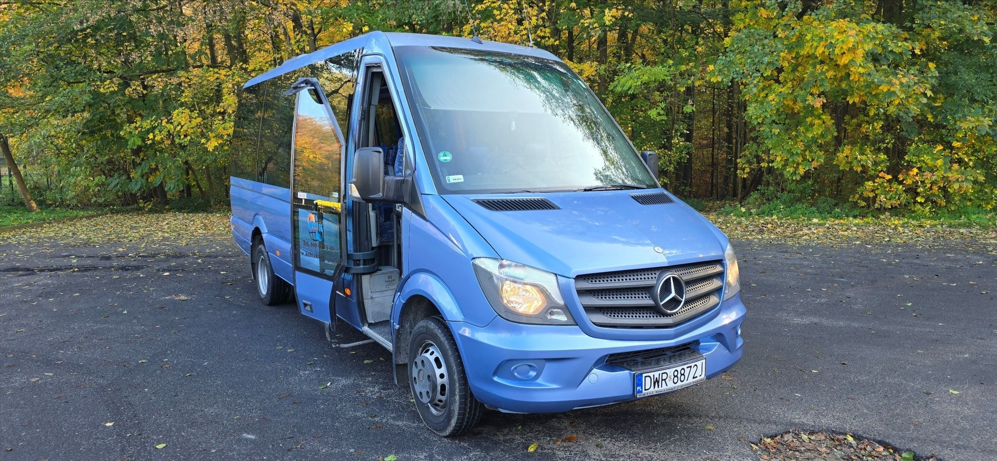 Niebieski bus Mercedes Sprinter z otwartymi drzwiami, zaparkowany na tle jesiennego lasu, widoczna tablica rejestracyjna.