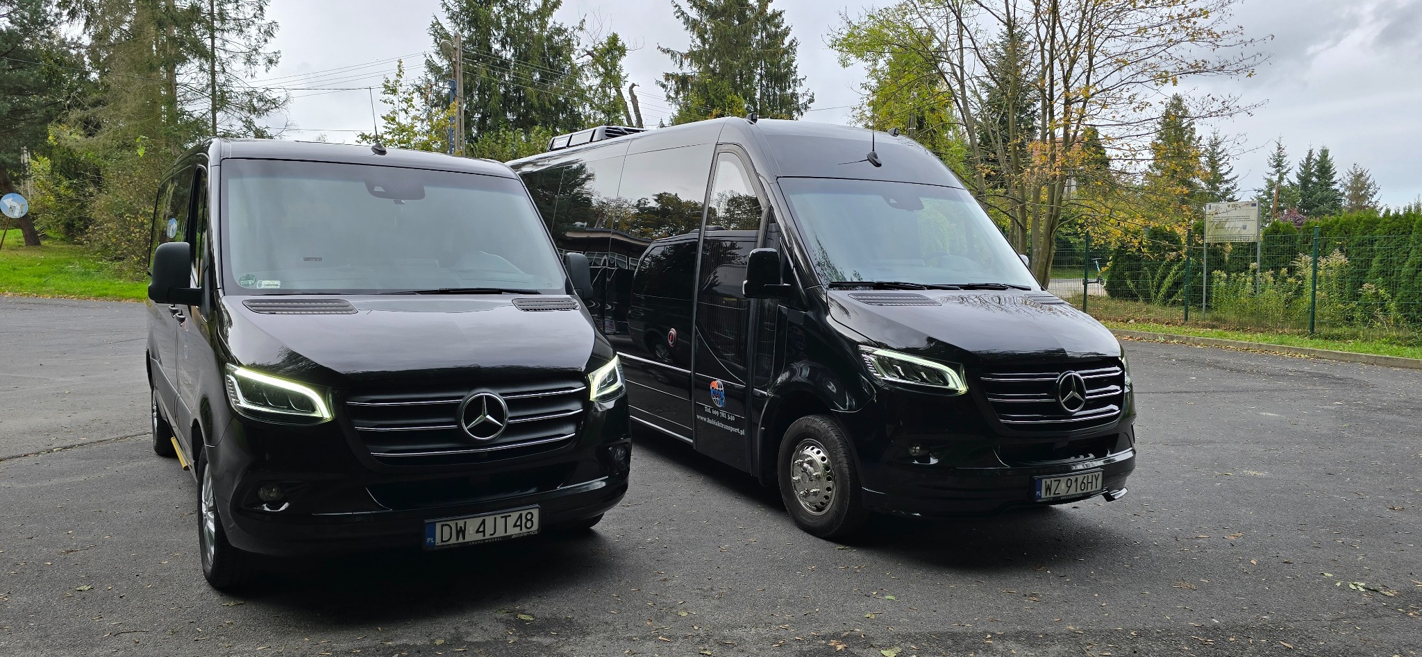Dwa czarne busy marki Mercedes-Benz Sprinter, jeden z bagażnikiem dachowym, zaparkowane na zewnątrz, widoczne tablice rejestracyjne.