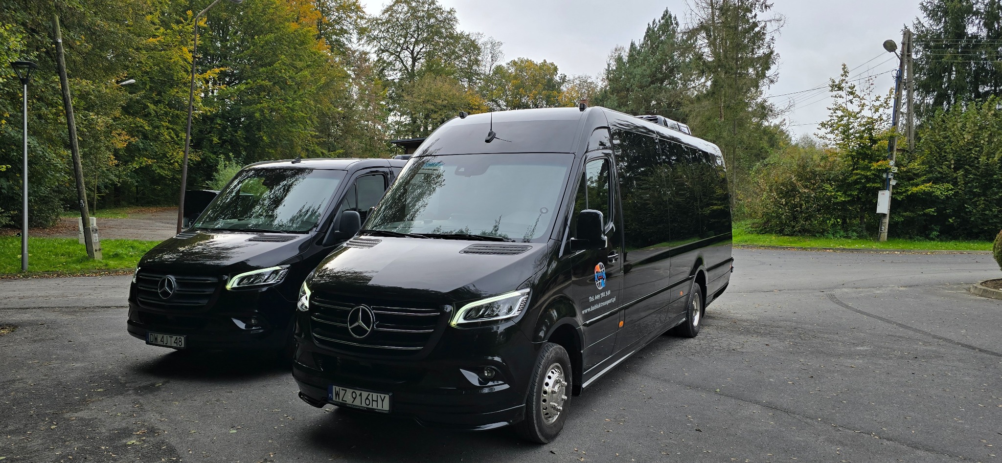 Dwa czarne busy Mercedes-Benz, jeden z widocznym logo firmy transportowej i numerem telefonu, zaparkowane na parkingu w otoczeniu drzew.